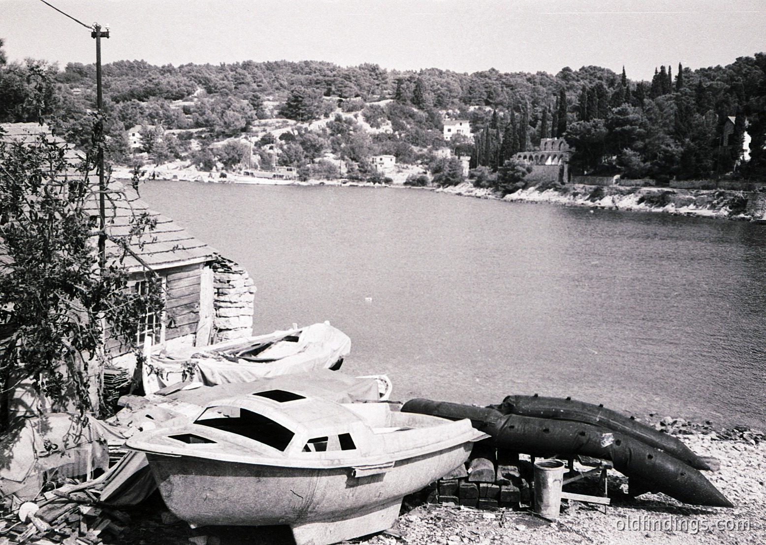 Mid-20th century coastal scene featuring a damaged wooden boat and WWII-era torpedo on a pebble shore. Dense forested hillside and scattered ruins in background suggest post-war Europe.