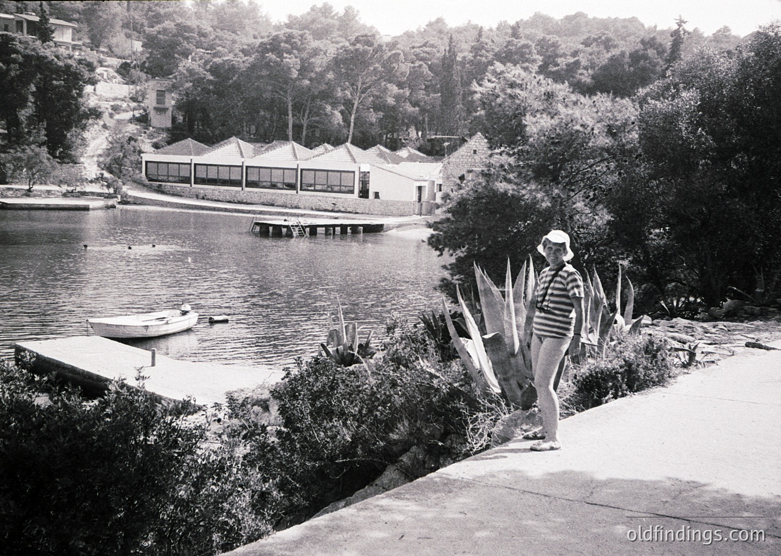 Mid-century lakeside resort with modernist pavilion and docked rowboat. Woman in striped swimsuit and hat stands near sun-bleached towels. Lush greenery and forested hills in background. Likely 1950s–1960s USA.