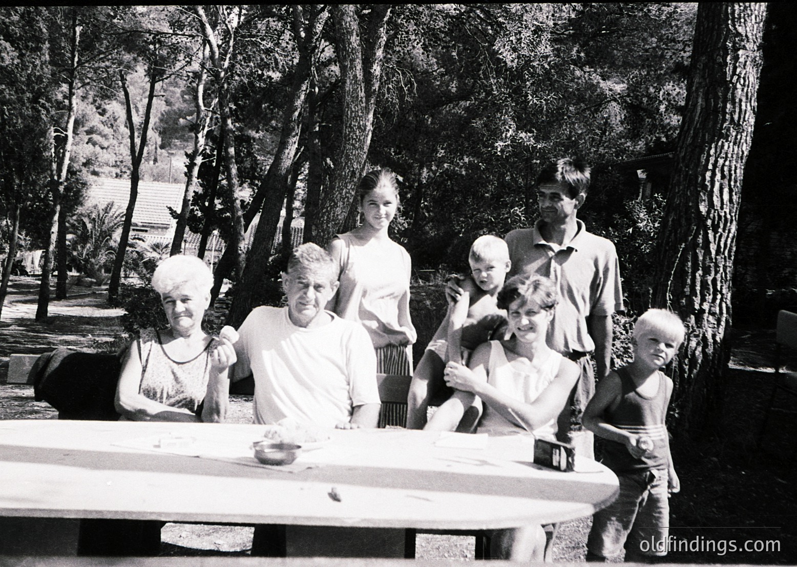Family portrait in a shaded outdoor setting, likely mid-20th century. Six individuals—two adults, three children, and an infant—pose around a wooden picnic table. Lush greenery and mature trees frame the scene, suggesting a park or garden. Clothing styles (short-sleeve shirts, dresses) and the black-and-white tone indicate a vintage aesthetic.