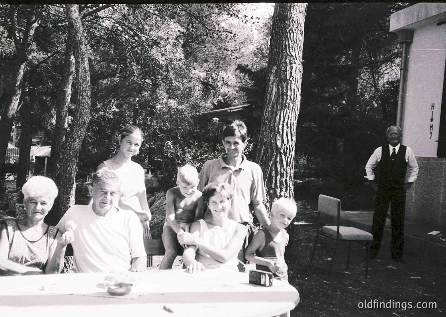 Family portrait in an outdoor setting, likely mid-20th century (1950s–1970s). Seven individuals pose around a picnic table under mature trees, with a man in a vest and tie standing to the right. Clothing suggests casual summer attire. Possible resort or park location.