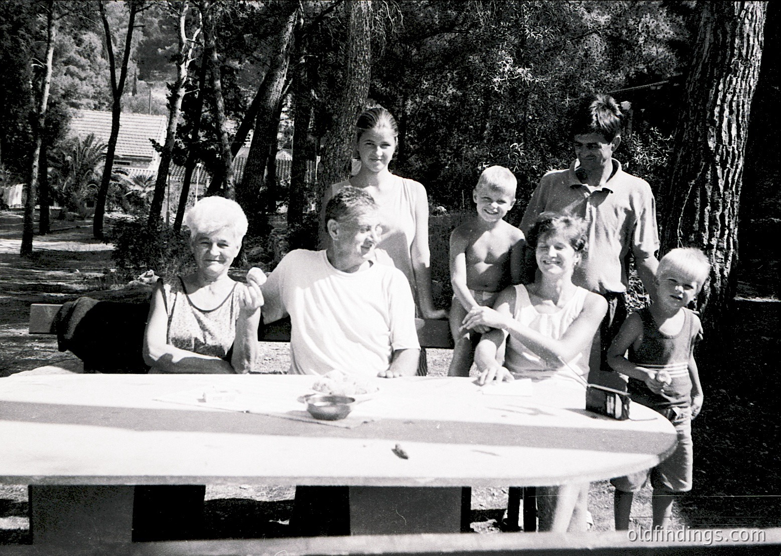 Family portrait in a lush, shaded outdoor setting, likely a resort or garden. Six adults and three children pose around a wooden table with a camera and small items. Mid-century clothing (1950s–1960s) suggests a post-war era. Tropical foliage and palm trees indicate a warm climate, possibly Mediterranean or Caribbean.