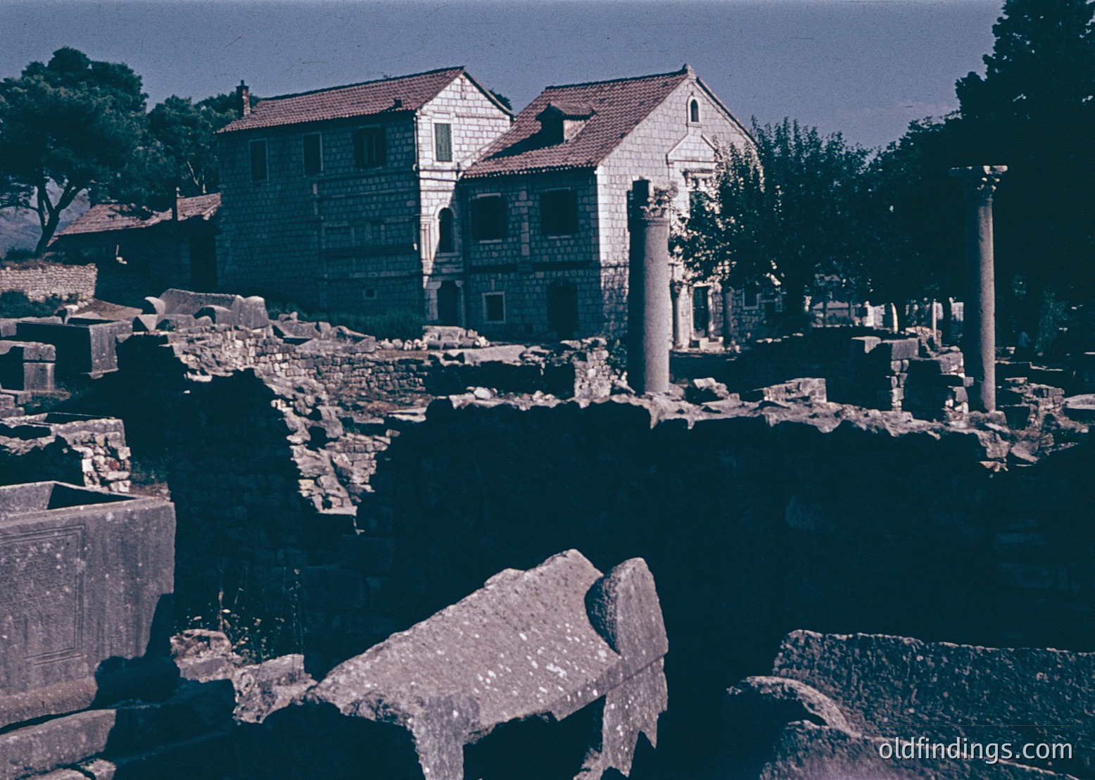 Blurred vintage photo of ancient ruins blending with modern structures—stone columns, fragmented marble, and a small water basin in foreground. Two-story building with tiled roof and decorative brickwork in background. Likely a preserved archaeological site in a Mediterranean climate.