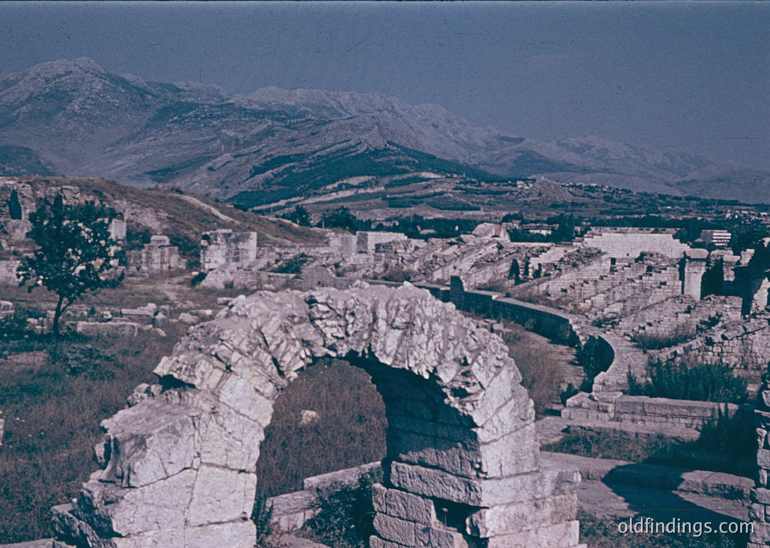 Ancient Roman triumphal arch fragment set against a mountainous landscape, likely part of a larger ruin complex. Mid-20th century black-and-white photo with slight color tinting.