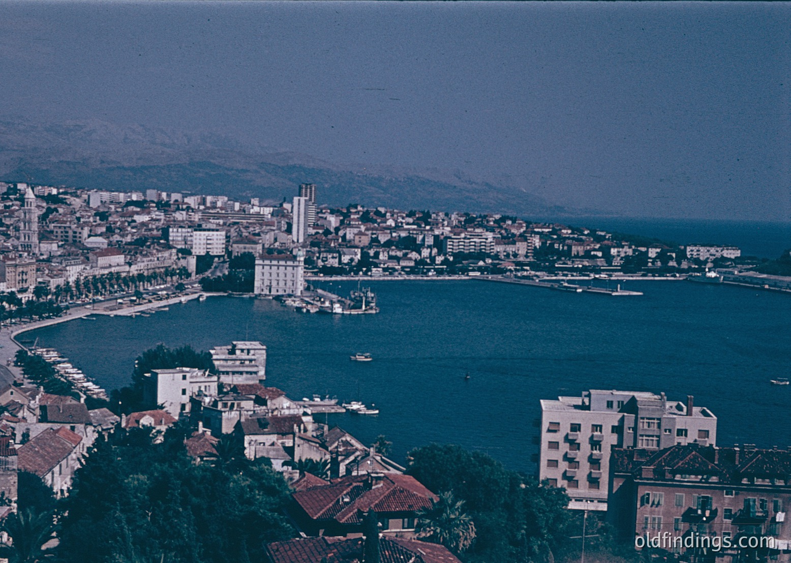 Aerial view of a coastal city with a prominent bay, featuring mid-20th century architecture. The scene includes a mix of low-rise buildings with red-tiled roofs and taller structures near the waterfront, likely from the 1960s-70s. The bay is bordered by a mix of urban and natural landscapes, with a notable church tower rising above the city. The coastline curves gently, with a visible pier extending into the water.