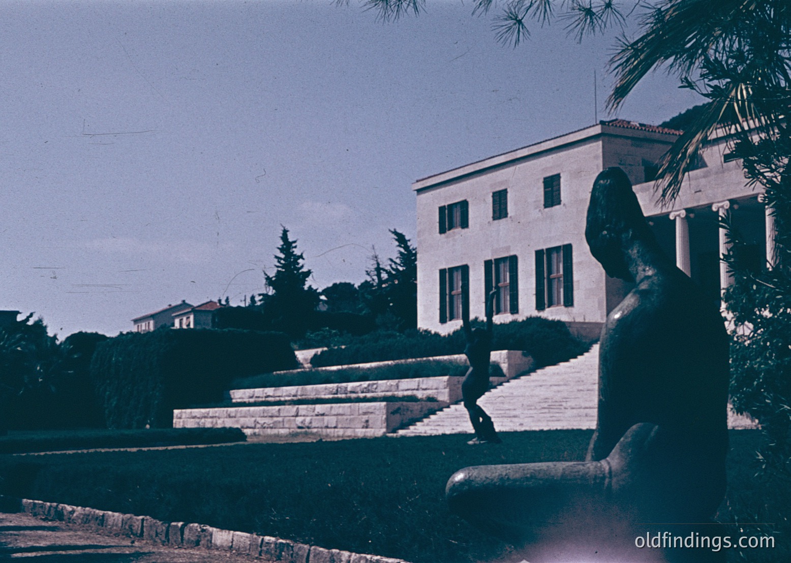 Vintage sepia-toned photo of a grand neoclassical building with symmetrical columns and large windows, likely a public institution. A statue of a seated figure dominates the foreground, positioned near a stone staircase. Lush greenery and a palm tree frame the composition, suggesting a Mediterranean or Southern European setting. Mid-20th century architectural style () with potential institutional or governmental use ().