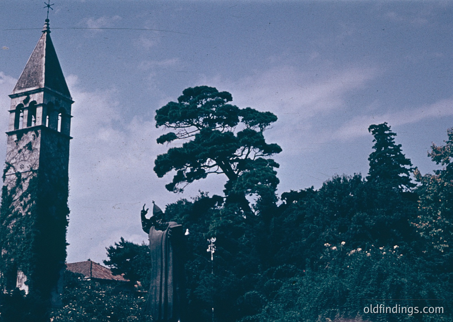 Vintage sepia-toned photo of a stone bell tower with arched openings, partially obscured by lush greenery. The tower’s weathered texture suggests aged masonry, likely from a European church or monastery. Dense foliage, including a prominent conifer, frames the scene, hinting at a rural or semi-rural setting. The sky is overcast, adding a muted, nostalgic tone.