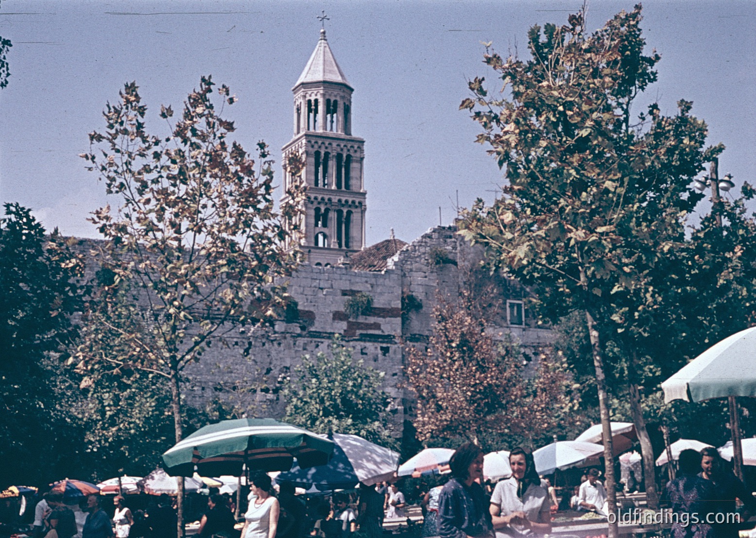 Historic bell tower with Romanesque arches dominates a bustling outdoor market in Split, Croatia. Stone architecture contrasts with vintage umbrellas and market stalls below. Likely 1960s–1970s based on color tone and attire.