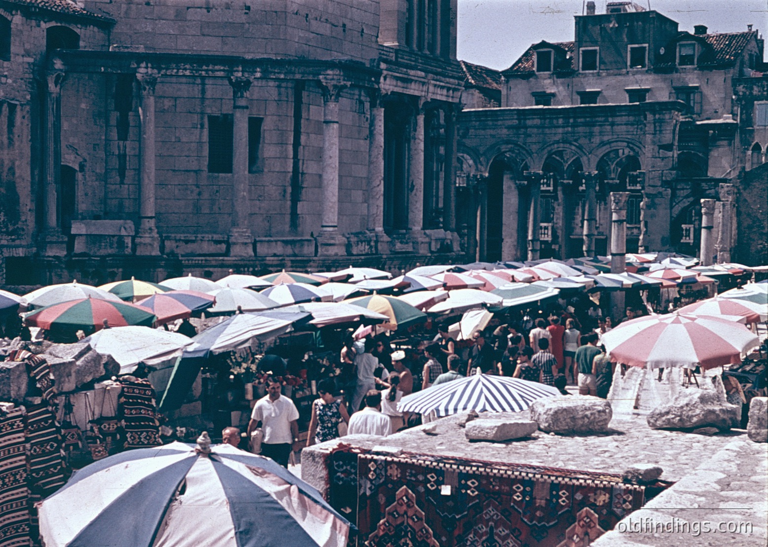 Vibrant 1960s/70s outdoor market in a historic European square, likely , Croatia. Roman-style arches and columns frame a bustling scene of striped umbrellas shielding vendors selling textiles, pottery, and souvenirs. Mid-century fashion and architecture blend in this lively public space.
