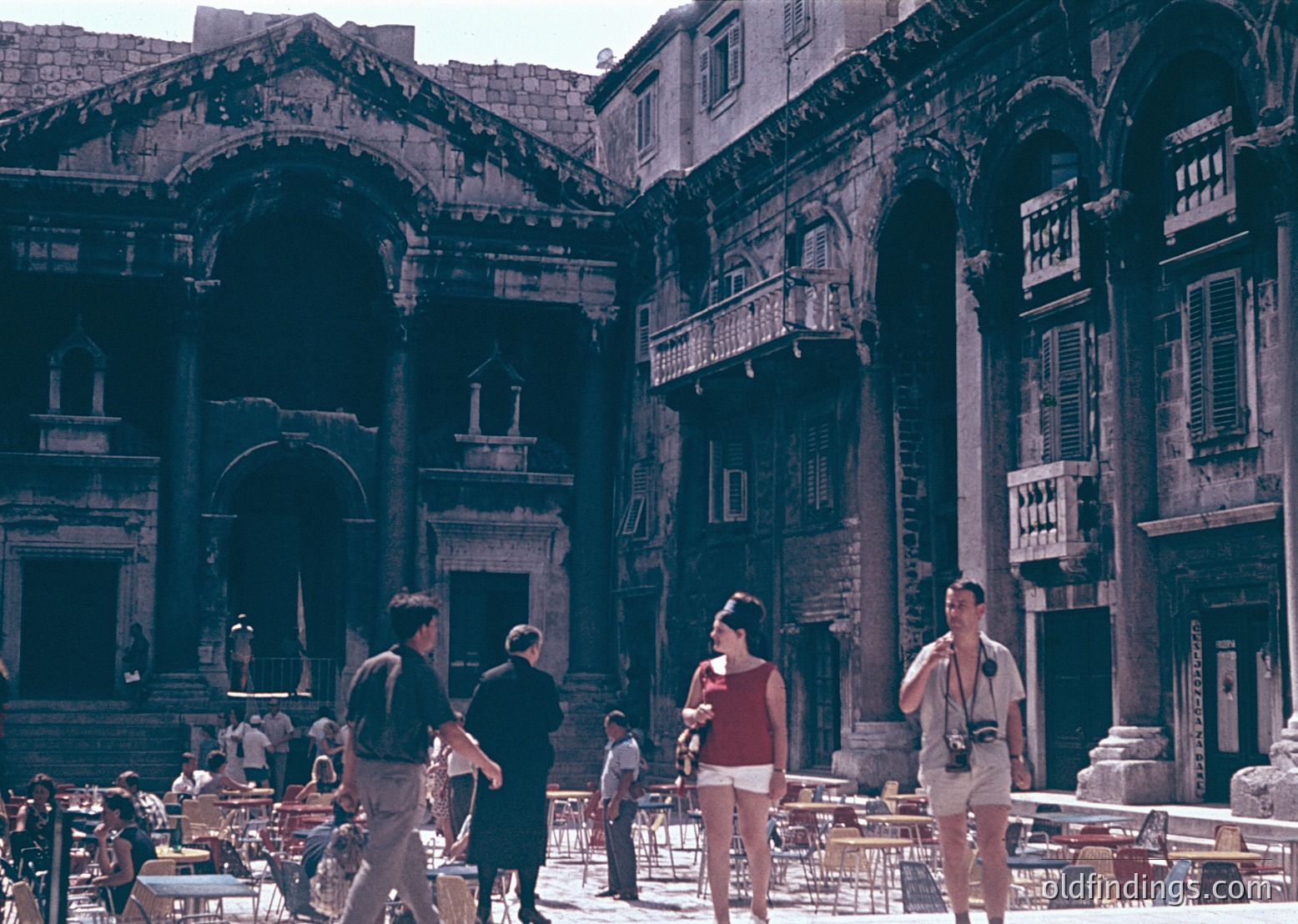 Roman Forum courtyard with ancient ruins and Renaissance-era balconies. Stone arches, columns, and weathered facades dominate the scene. Mid-20th-century tourists in casual attire (1950s–60s) explore the historic site. Outdoor café tables and chairs suggest a blend of ancient and modern activity.