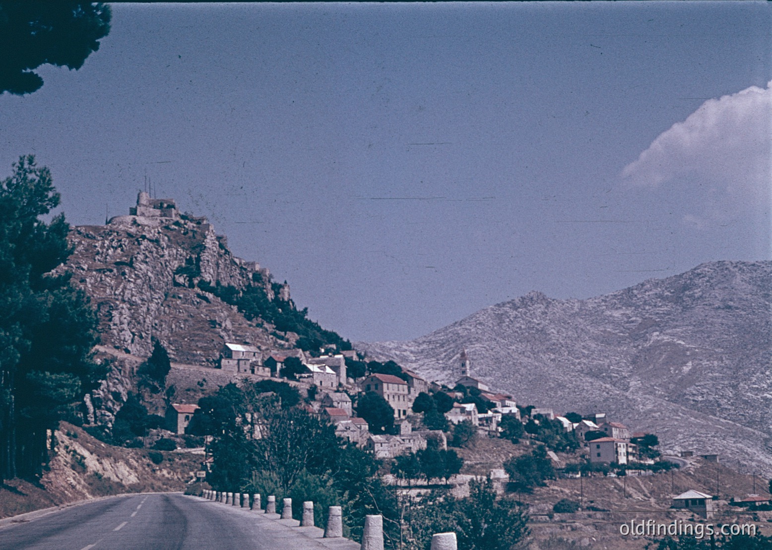 Vintage aerial-style view of a Mediterranean hillside village with terracotta-roofed houses nestled among rugged terrain. A winding road curves through the foreground, flanked by concrete guardrails. Distinctive watchtower perched atop a rocky outcrop. Likely –1970s