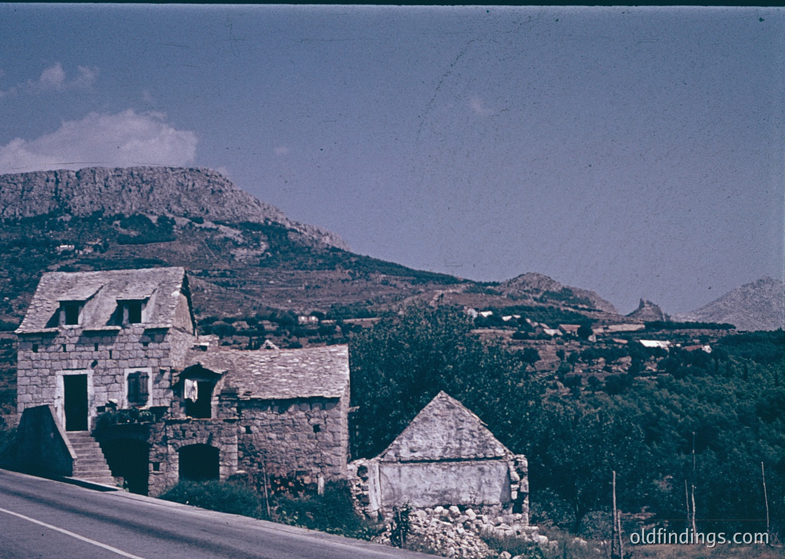 Vintage stone farmhouse cluster nestled in a Mediterranean hillside landscape, likely -70s. Rustic architecture with flat roofs, arched doorways, and weathered walls contrasts against rugged terrain and distant peaks. *(Note: Specific location indeterminate but stylistically aligned with Mediterranean/Southern European regions.)*