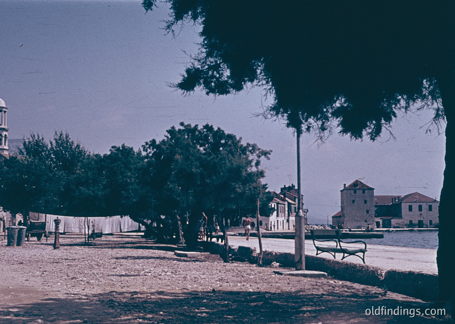Vintage seaside promenade with mid-20th century architecture. A row of trees lines a paved walkway beside a body of water, with laundry hung on a line in the foreground. Distant buildings—likely residential or small hotels—feature flat roofs and simple facades. A lone wrought-iron bench sits near the water’s edge.