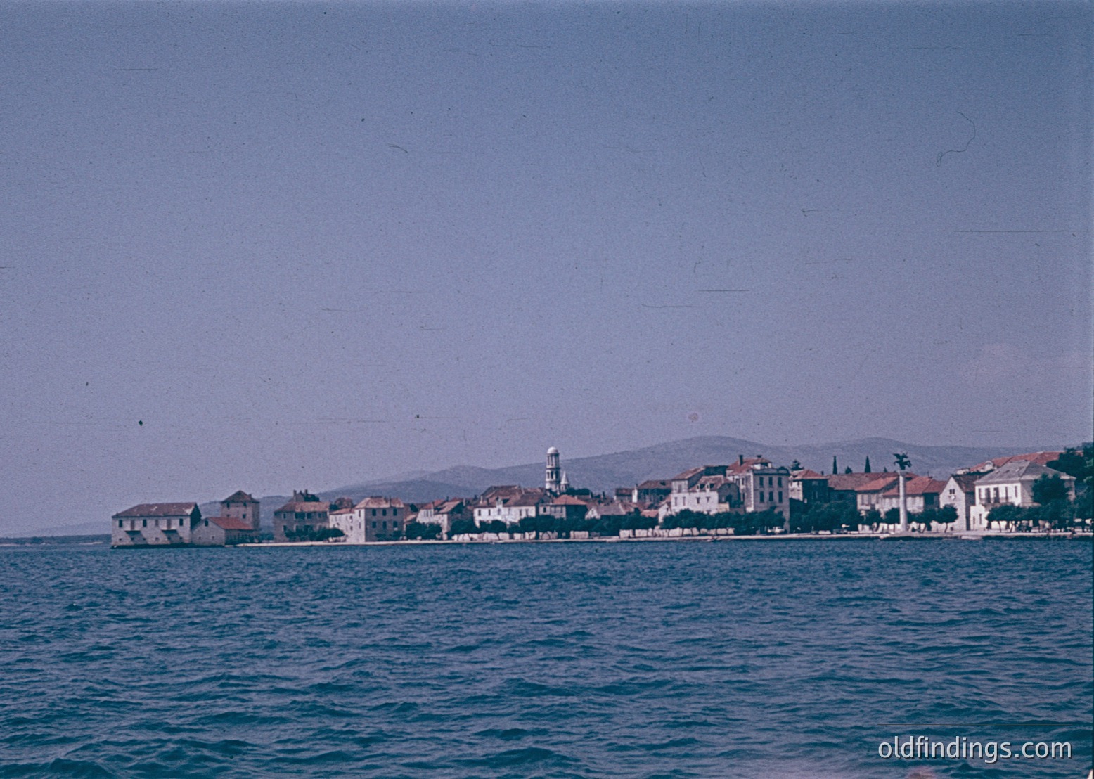 Vintage coastal town with low-rise buildings and a prominent lighthouse on a rocky outcrop. Waterfront architecture features flat roofs and light-colored facades. Likely Mediterranean or Adriatic region, mid-20th century.