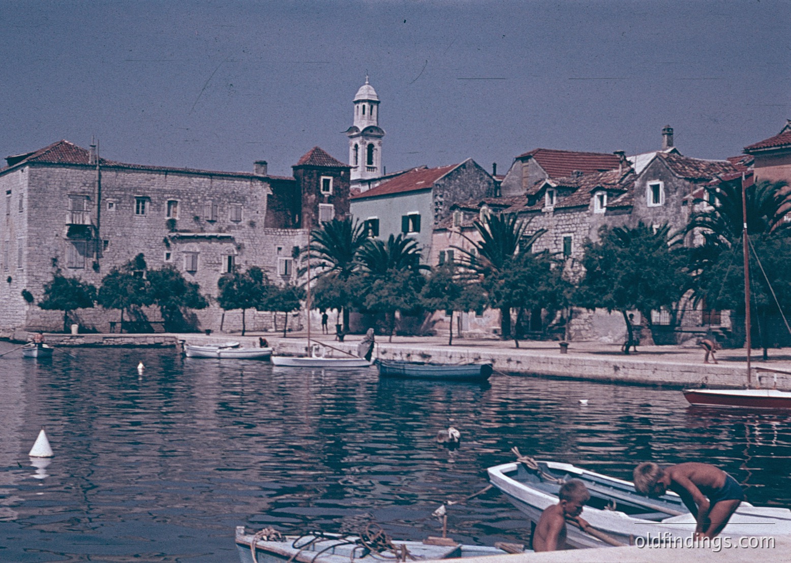 Vintage coastal scene featuring a historic stone fortress by the waterfront, likely Mediterranean. Two men in swim trunks work on a small boat in the foreground, while palm trees and traditional architecture line the shore. The prominent bell tower suggests a church or municipal building. Likely **1950s-1960s**, **Croatia** ().