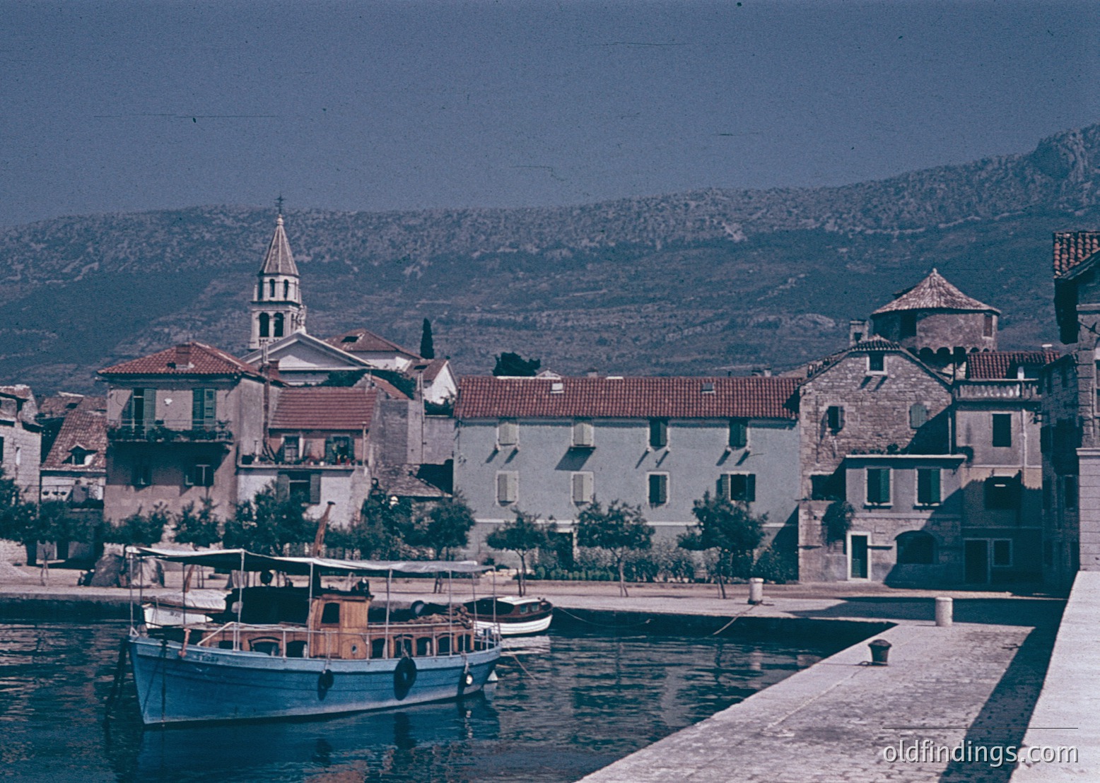 Vintage coastal town with Mediterranean architecture—stone buildings, terracotta roofs, and a prominent church steeple. Small wooden boats docked along a calm waterfront, framed by lush greenery and rolling hills. Likely Adriatic or Aegean region, 1960s–1970s era.