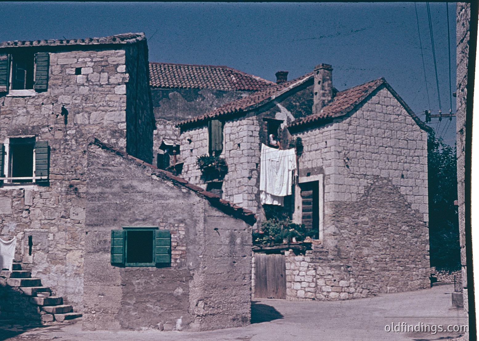 Stone-built Mediterranean village with terracotta-tiled roofs, likely –70s. Clothes drying on a balcony and window ledge suggest domestic daily life. Rustic wooden shutters and narrow alleyways reflect traditional architecture. Potential research value for rural European heritage studies.