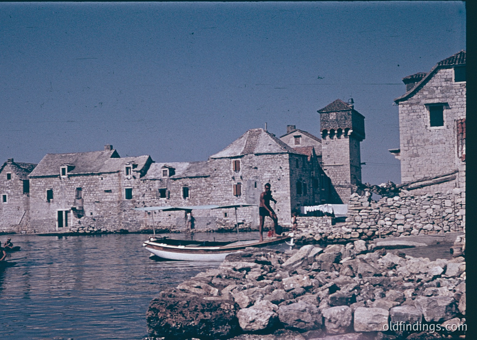 Historic stone fortress by waterfront, featuring medieval towers and low-rise buildings. A man in traditional attire stands near a small wooden boat. Likely Dalmatian coastal region, architecture. --- **Notes:** - **Visual content:** Stone fortress walls, towers, low-rise buildings, wooden boat, man in traditional attire. - **Context:** Likely Dalmatian region (Croatia), given architectural style and coastal setting. - **Time period:** Mid-20th century (1960s), inferred from clothing and photo grain. - **Commercial value:** Useful for historical research, stock photography of coastal heritage, or design references for Mediterranean-inspired projects.