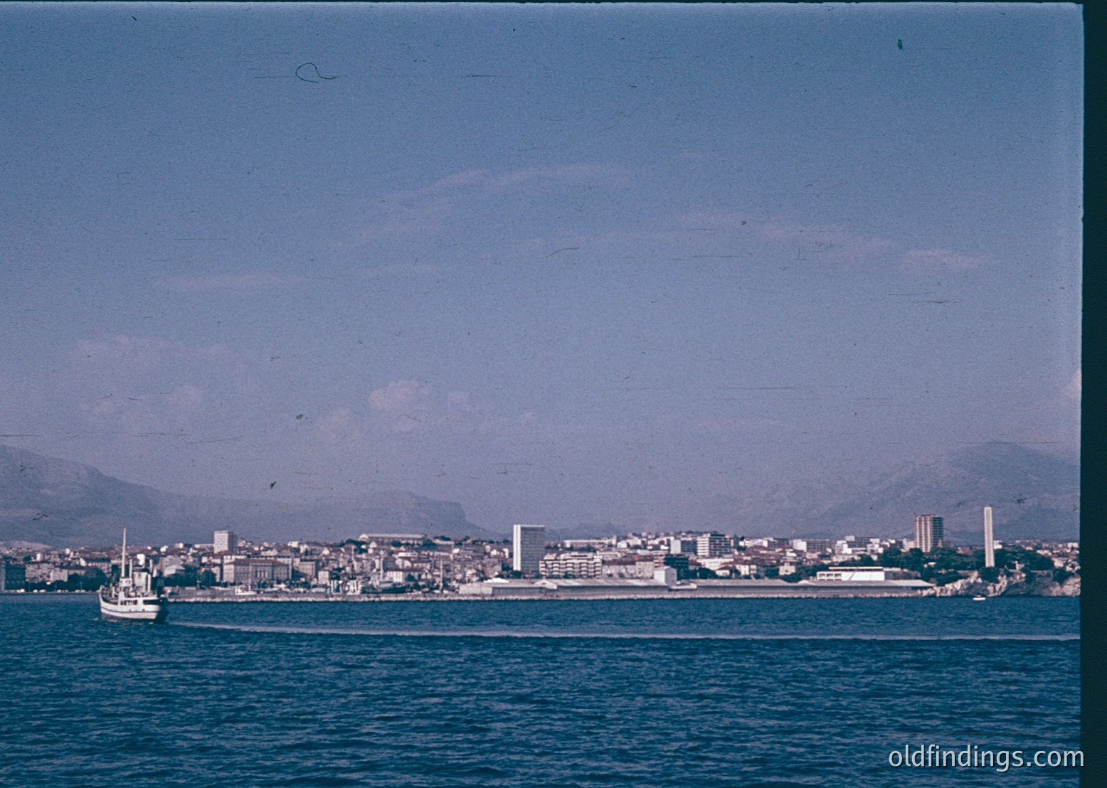 Vintage coastal cityscape with low-lying buildings, likely a Mediterranean port. A small boat floats in the foreground, while a distant mountain range frames the horizon. Mid-20th century architecture suggests midcentury urban development. Waterfront infrastructure hints at maritime trade or tourism.