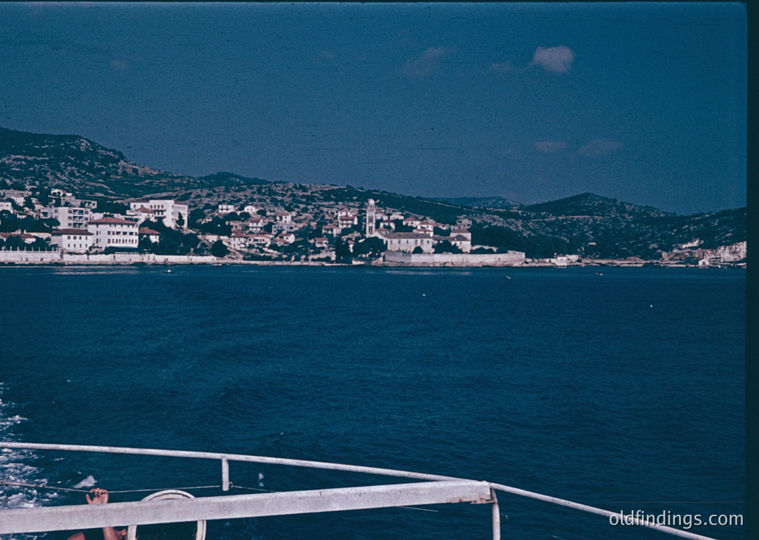 Coastal town nestled between deep blue waters and rolling hills, featuring clustered whitewashed buildings with red-tiled roofs. Prominent church tower and lush greenery dominate the hillside. Likely Mediterranean, 1960s–1980s era.