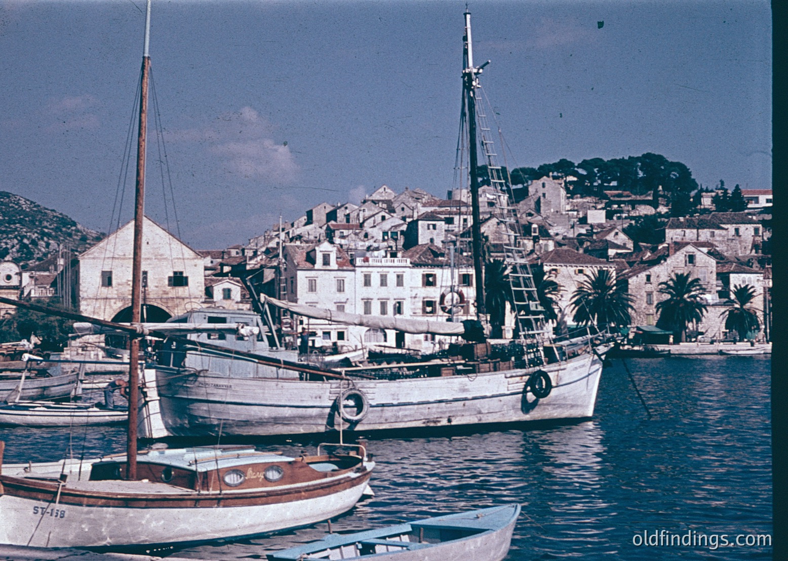 Classic wooden sailing vessel docked in a Mediterranean coastal town, mid-20th century. Whitewashed buildings with terracotta roofs and palm trees line the waterfront. Vintage color tones suggest 1950s–1960s era.
