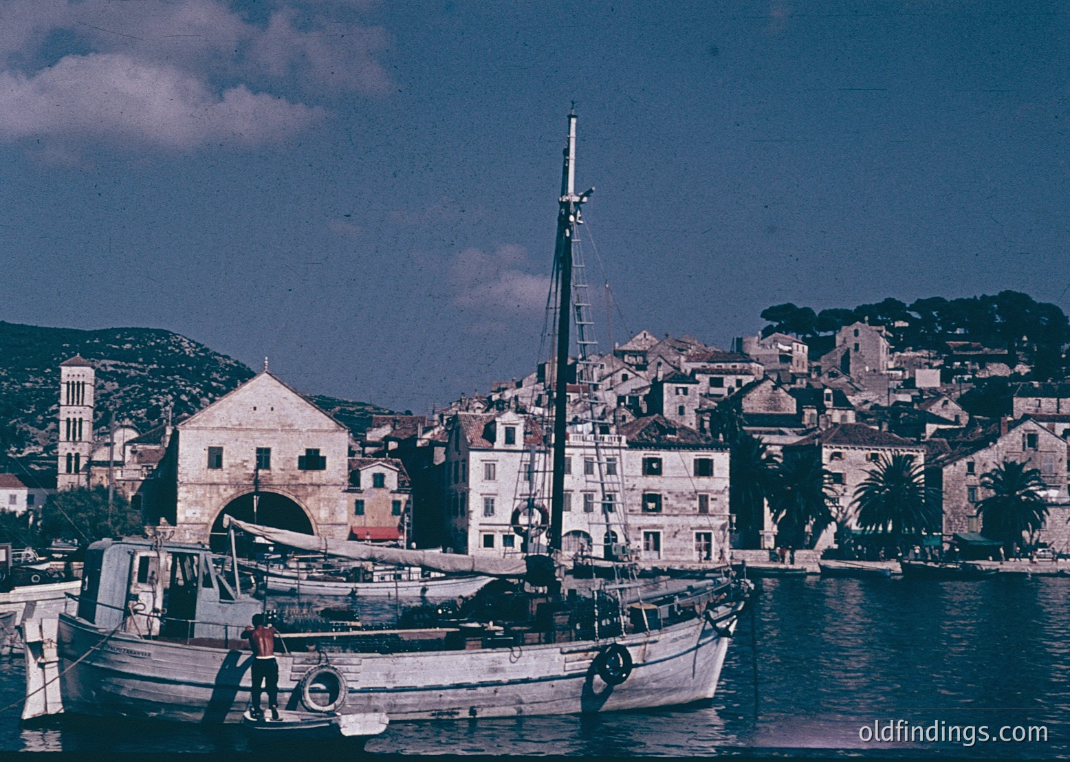 Vintage coastal scene featuring a small wooden fishing boat docked near a historic Mediterranean harbor. Whitewashed buildings with arched doorways and terracotta roofs line the waterfront, framed by lush greenery and rolling hills. The photograph’s sepia tone suggests 1950s–1960s era.
