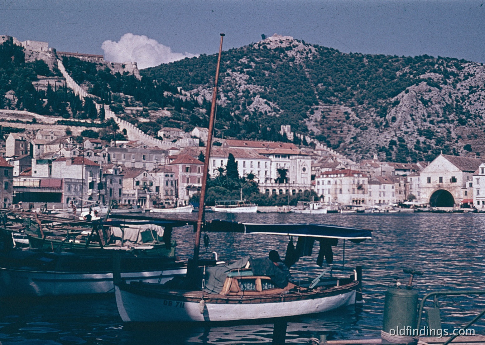 Historic coastal town with Mediterranean architecture, featuring whitewashed buildings and a prominent fortress on rocky hills. Small wooden boats docked in a harbor, likely from the 1960s–70s. Clear waters reflect the vibrant urban landscape.
