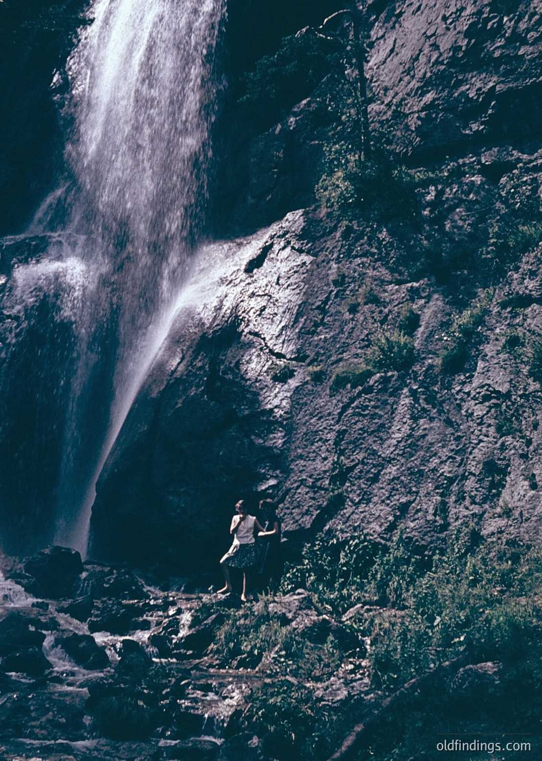 Black-and-white shot of a lone figure standing near a powerful waterfall cascading down rocky terrain. Dramatic lighting highlights mist and texture.