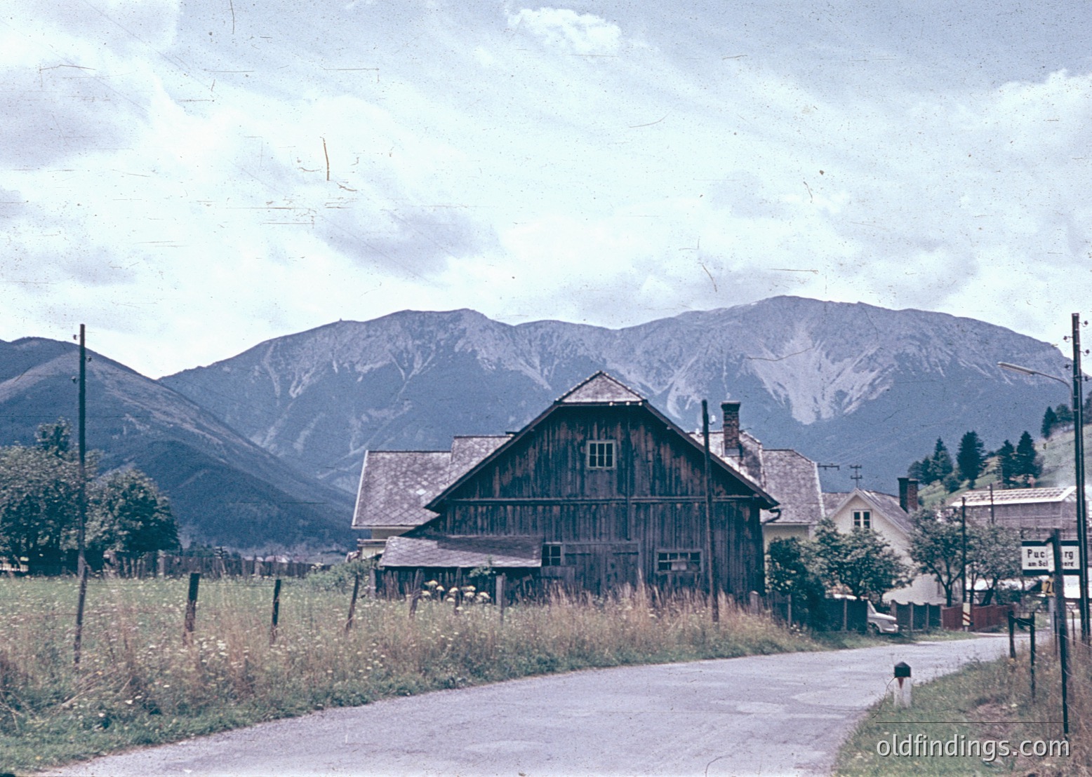 Vintage alpine village scene featuring a rustic timber-framed barn with steep gable roof, set against snow-capped mountains. Mid-20th century rural architecture with unpaved road and sparse vegetation. Likely European alpine region.
