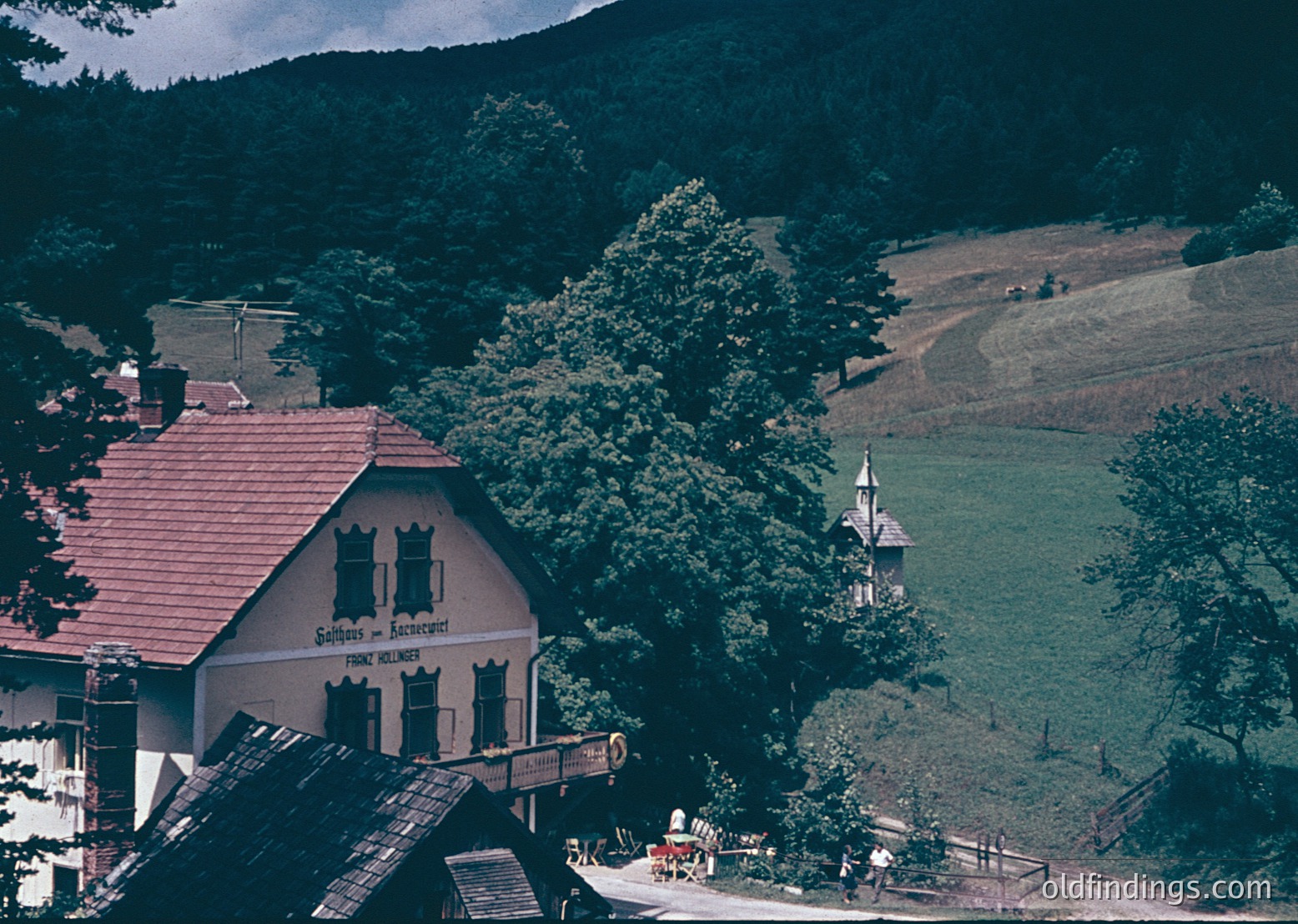 Vintage alpine lodge with red-tiled roof and sign reading *"Gasthof & Alpenrestaurant"* (est. 1898), nestled among dense forest. Rolling green meadows and distant church steeple in hilly countryside. Likely European alpine region, mid-20th century.