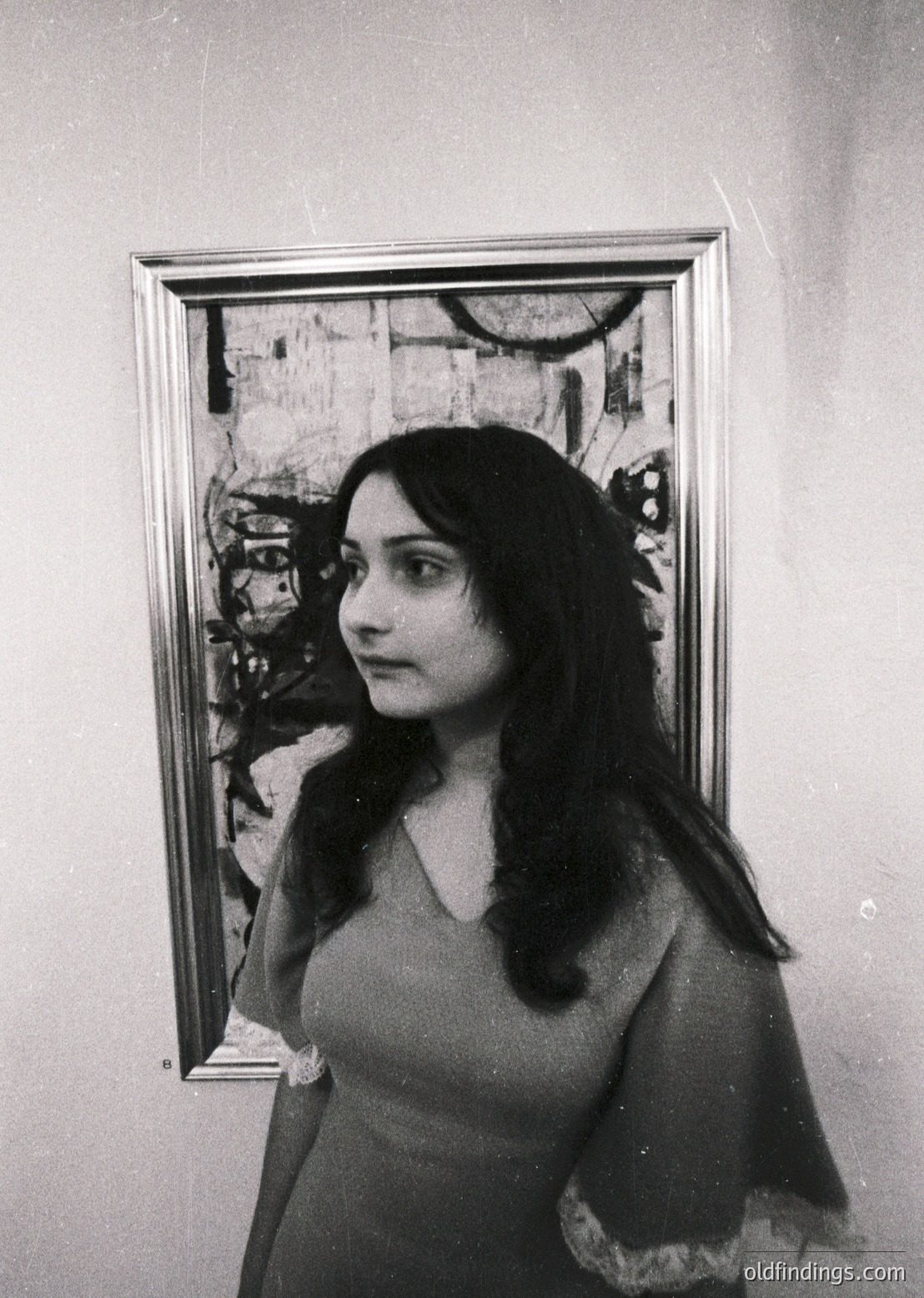 Portrait of a woman in a sleeveless dress, standing beside an abstract painting with bold, angular forms. The composition suggests a mid-20th-century art gallery or studio setting, likely or . The woman’s hairstyle and clothing reflect trends of the era.