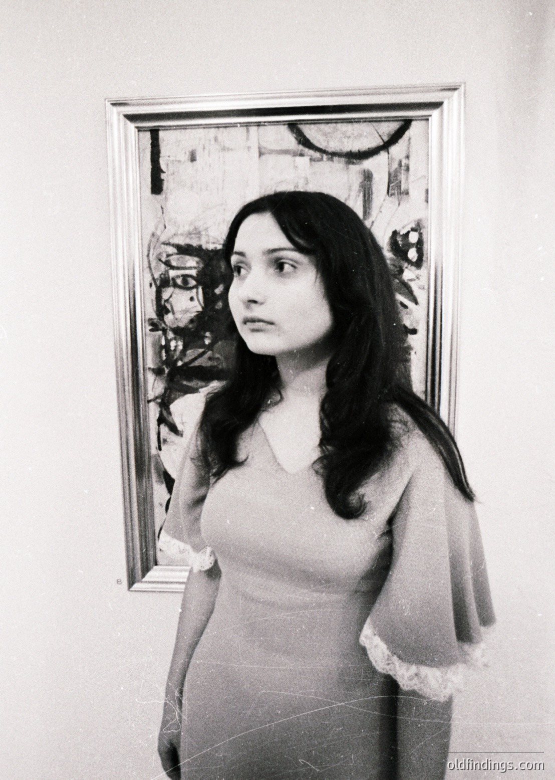 Portrait of a woman in a 1960s–70s abstract art gallery, framed by an expressive modernist painting. She wears a sleeveless dress with lace trim, posed against a minimalist white wall.