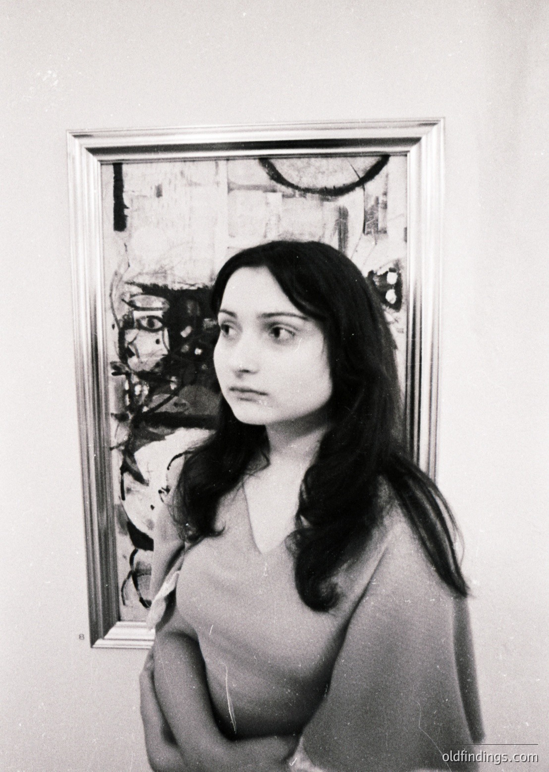 Portrait of a woman in a sleeveless dress, standing beside an abstract painting with bold, gestural brushstrokes and muted tones. The composition suggests an art gallery or studio setting, likely mid-20th century. The woman’s pose and attire reflect a timeless elegance.