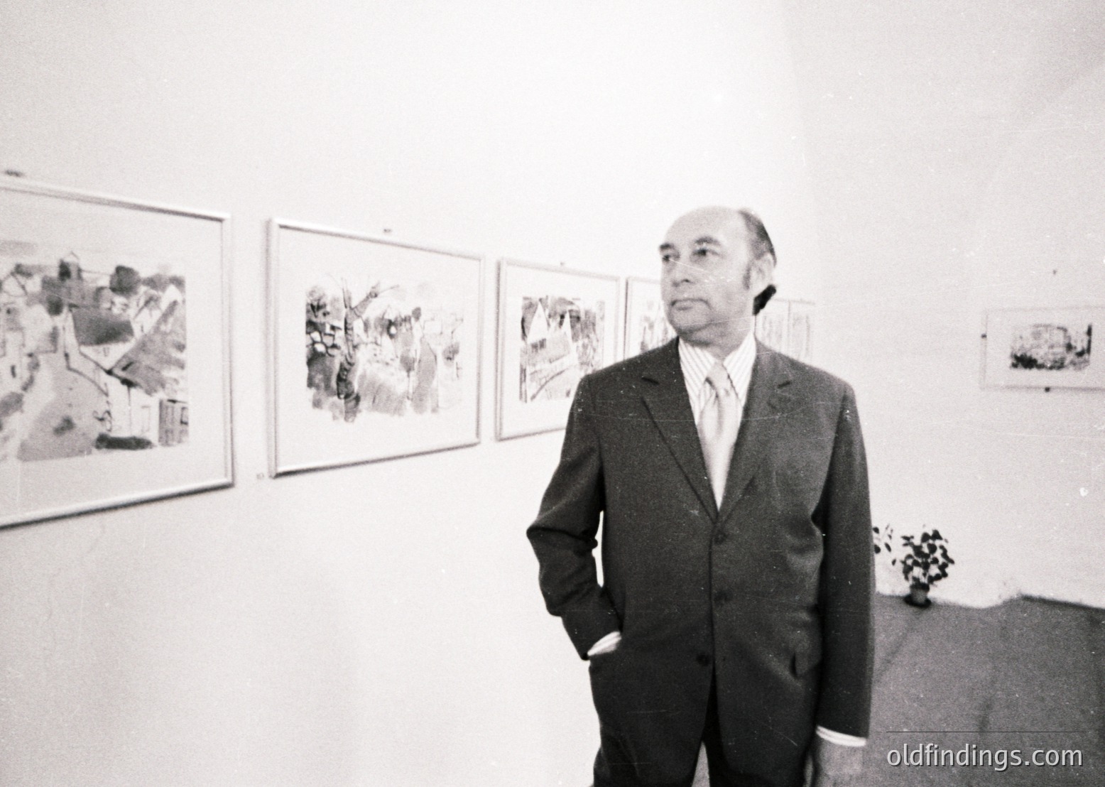 Mid-century man in a dark suit poses in a gallery, surrounded by abstract architectural sketches. Clean lines and minimalist framing suggest 1960s–70s modernist influence. Potential reference for design history or architecture studies.