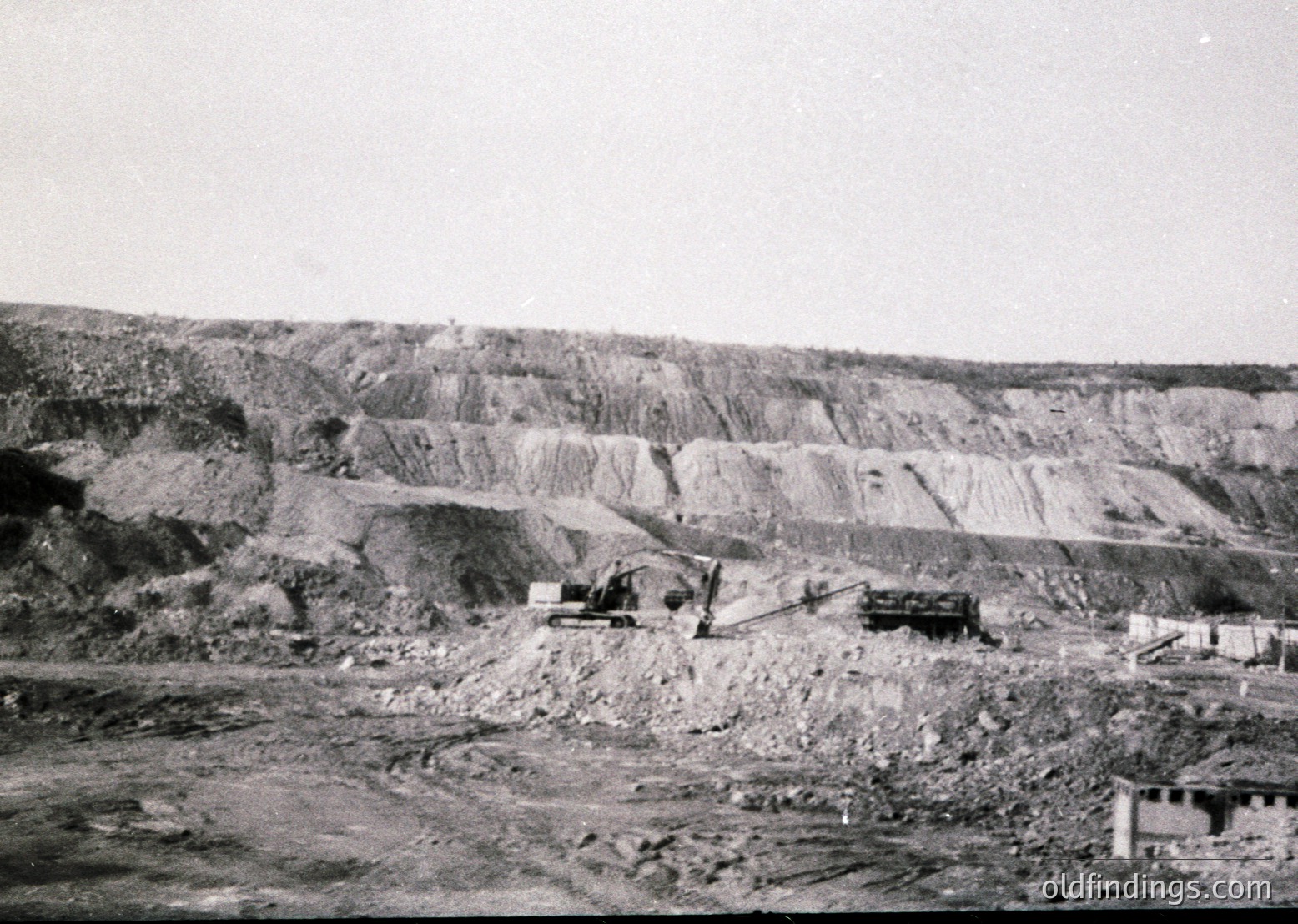 Open-pit mining site with layered rock strata and heavy machinery—likely a bulldozer and conveyor system. Mid-20th century industrial landscape, suggesting resource extraction.