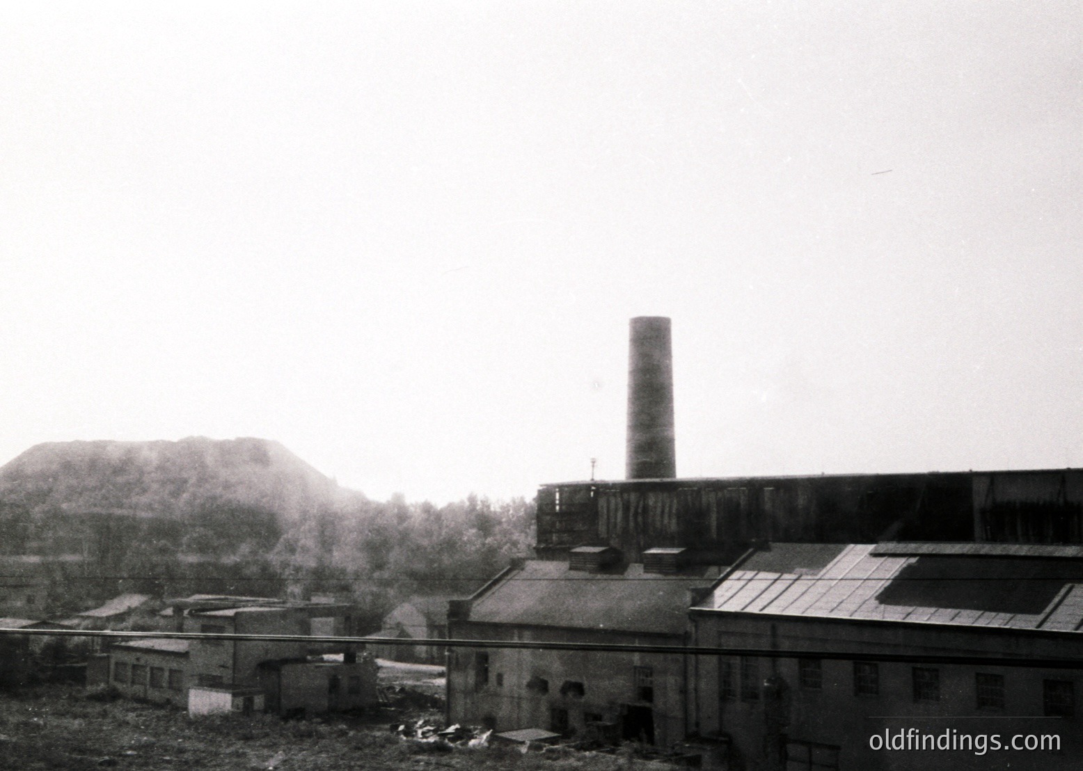 Industrial complex featuring a tall brick smokestack and multi-story factory buildings, likely mid-20th century. Overcast skies and misty terrain suggest a damp, possibly Eastern European setting. Raw, utilitarian architecture reflects post-war industrialization.