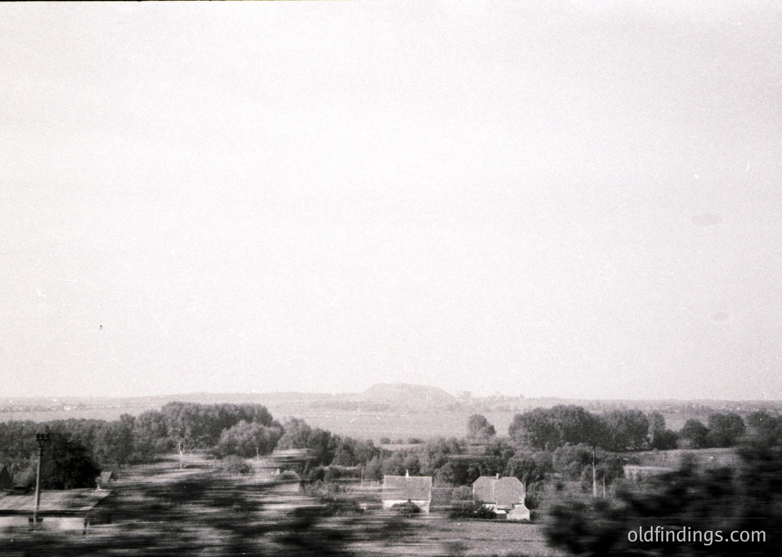 Vintage black-and-white rural landscape featuring scattered farmhouses, dense tree clusters, and open fields stretching to a distant hill. Likely Eastern European countryside, mid-20th century.