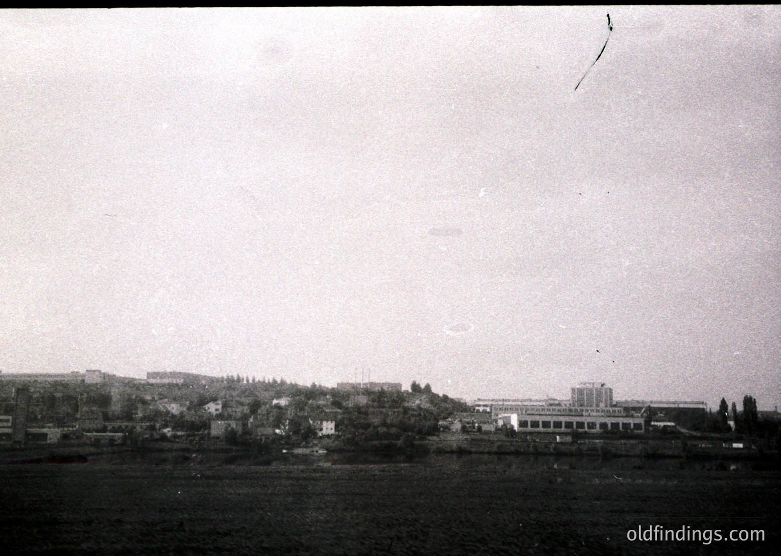 Vintage black-and-white coastal industrial scene featuring a long, low-lying building complex along a waterfront, likely a factory or port facility. Dense tree line separates structures from the shoreline. Overcast sky enhances moody, historical atmosphere. Potential Eastern Bloc-era architecture () suggests Soviet-era industrial design.
