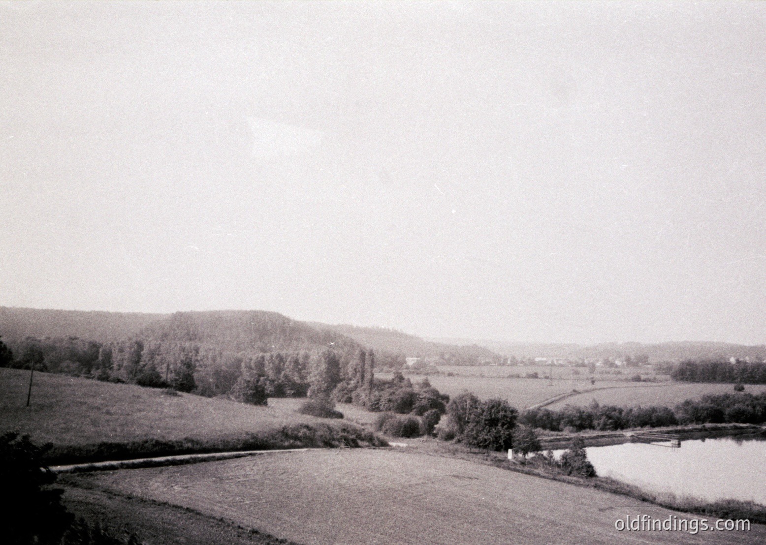Vintage black-and-white rural landscape featuring a winding road bordered by dense forest on one side and open fields on the other. A small body of water, likely a pond or river, curves gently through the foreground. Mid-20th century agricultural setting with sparse structures visible in the distance.