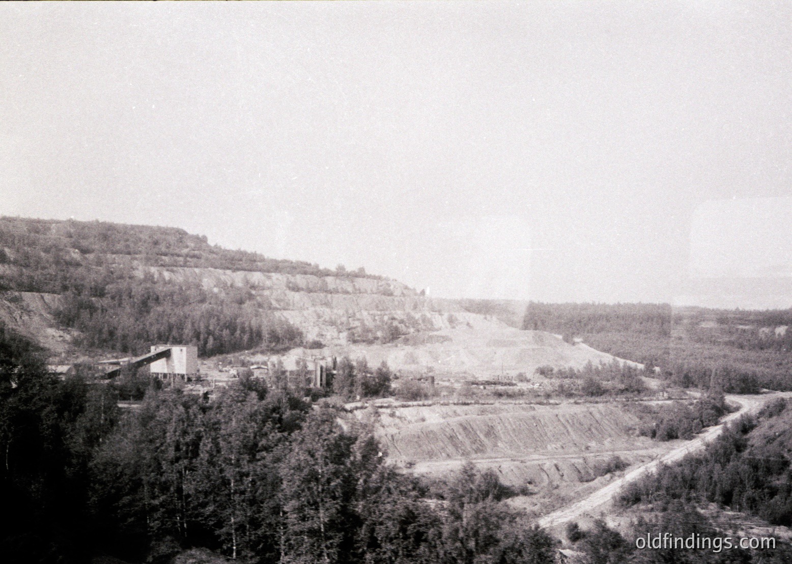 Mid-20th century industrial landscape featuring an open-pit mine with terraced excavation. Dense forest surrounds the site, contrasting with the exposed earth. Low-lying industrial structures and a road network suggest mining operations. Likely Eastern European due to architectural style and vegetation.