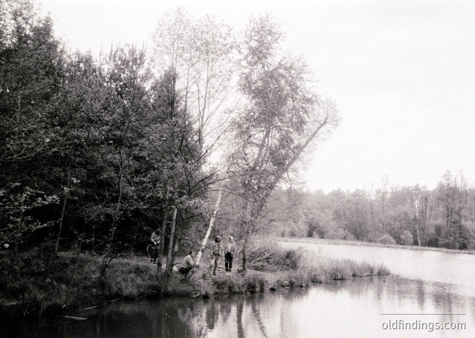 Black-and-white shot of three figures standing near a shallow lake edge, surrounded by dense forest. Reflective water and overcast sky enhance serene atmosphere. Likely mid-20th century outdoor recreation scene.