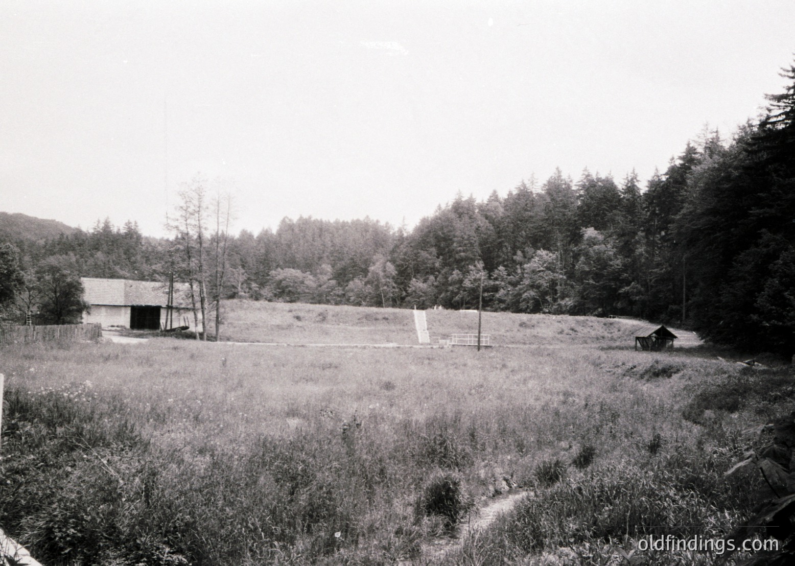 Black-and-white rural landscape featuring a small wooden barn and shed in a grassy clearing. Dense forest surrounds, with a dirt path leading through the scene. Likely early-to-mid 20th century, suggesting historical farmstead or countryside setting.