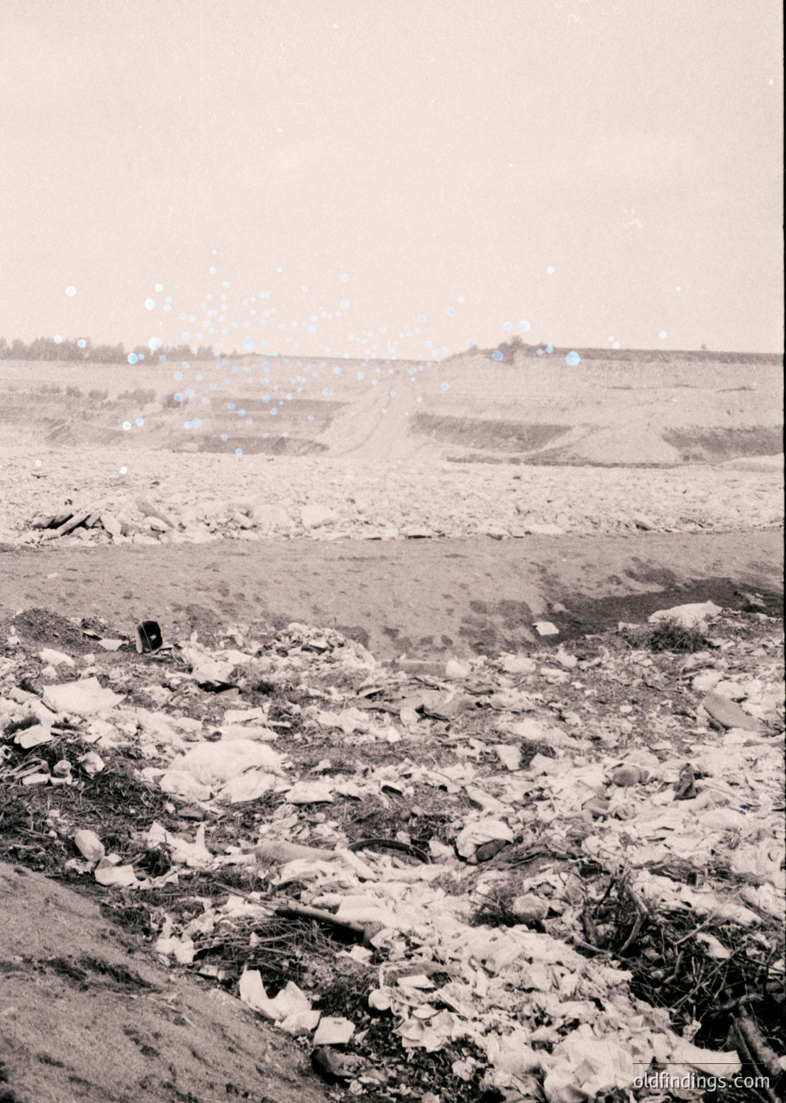 Barren, snow-dusted landscape with scattered debris and sparse vegetation, likely post-industrial or abandoned. Distant low-lying structures hint at human presence. Monochrome suggests vintage or archival quality. Potential or era.