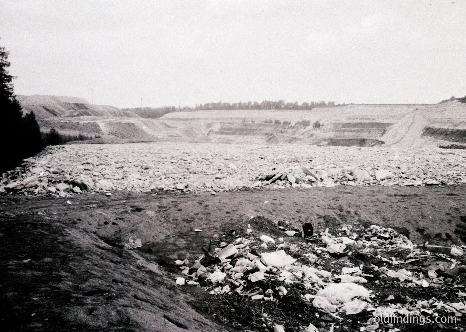 Black-and-white industrial site featuring layered rock excavation, likely limestone or marble quarry. Foreground shows loose debris and partially processed blocks. Midground reveals terraced excavation pits with visible horizontal strata. Background includes sparse forest and distant infrastructure. Suggests mid-20th century mining activity.