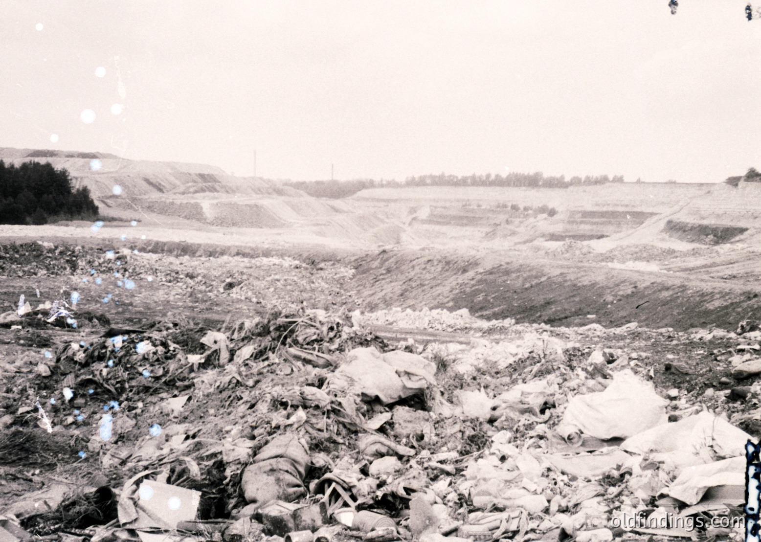 Mid-20th century industrial waste site with discarded materials including glass, metal, and debris. Open-pit excavation in background suggests mining or quarrying activity.