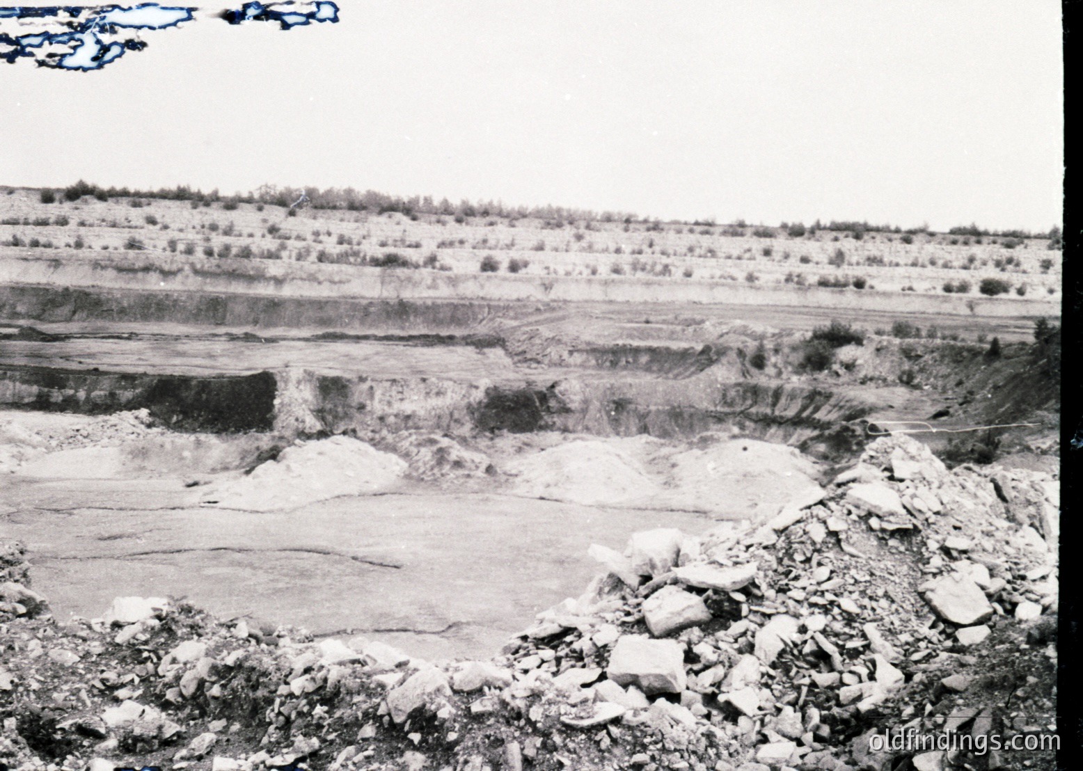 Mid-20th century open-pit mining site with exposed stratified rock layers, likely limestone or gypsum. Foreground shows loose rock debris and vegetation. Background reveals terraced excavation with minimal industrial equipment. Landscape suggests arid or semi-arid climate.