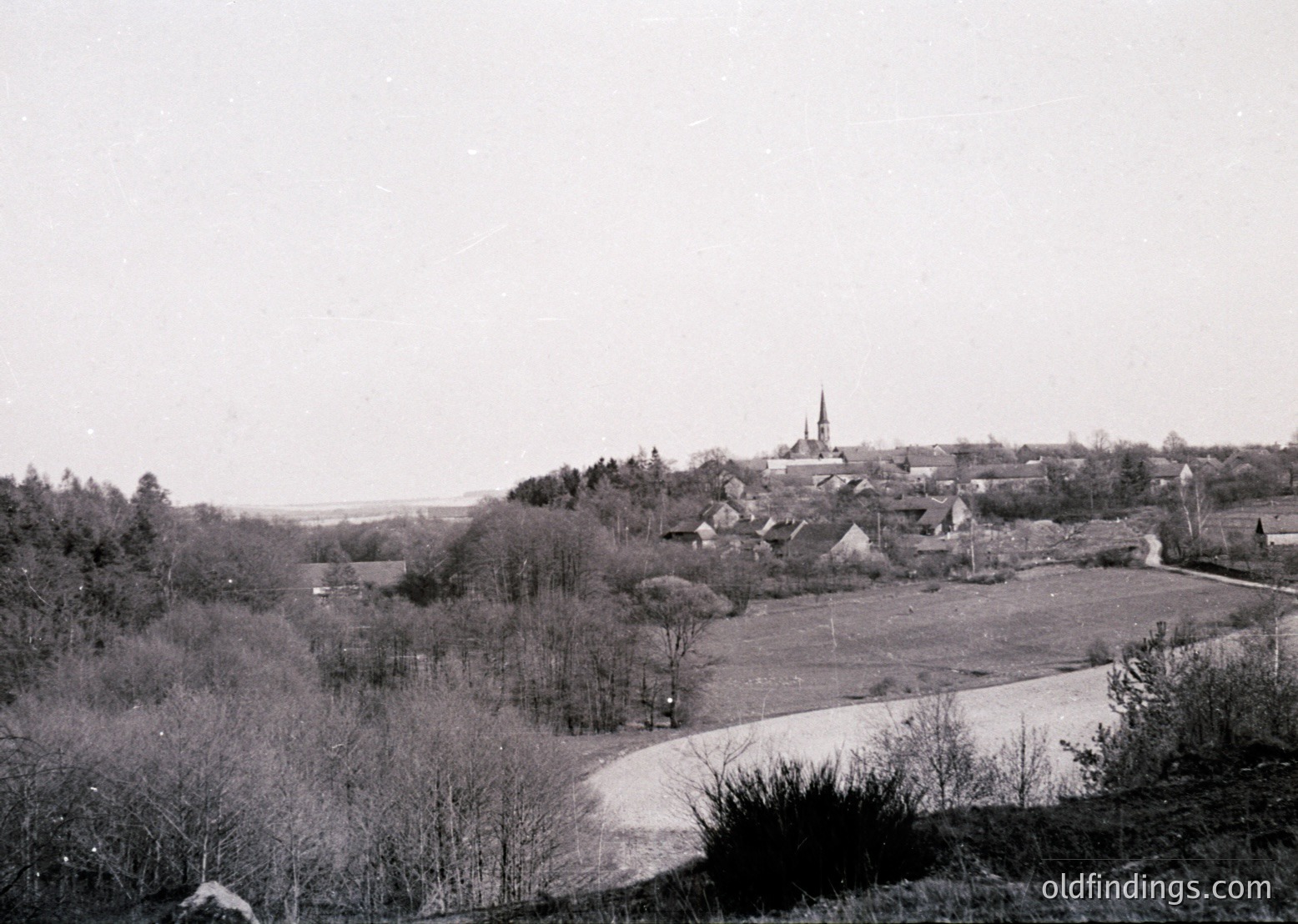 Vintage black-and-white rural village scene with a prominent church steeple and scattered wooden houses. Open fields and leafless trees suggest late winter or early spring. Road curves gently through the landscape, hinting at early 20th-century European countryside.