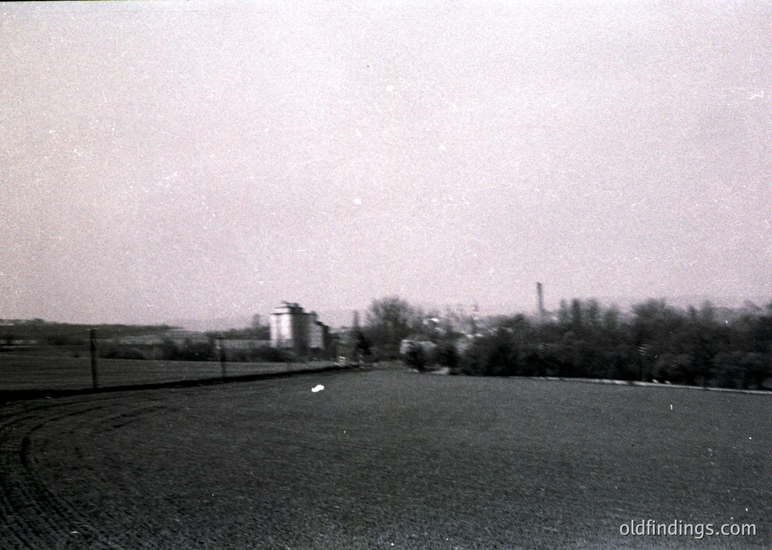 Mid-20th century industrial landscape with blurred motion, likely from handheld camera. Foreground shows a winding rural road bordered by a fence, leading toward a smokestack and factory complex. Dense tree line separates foreground from industrial area.
