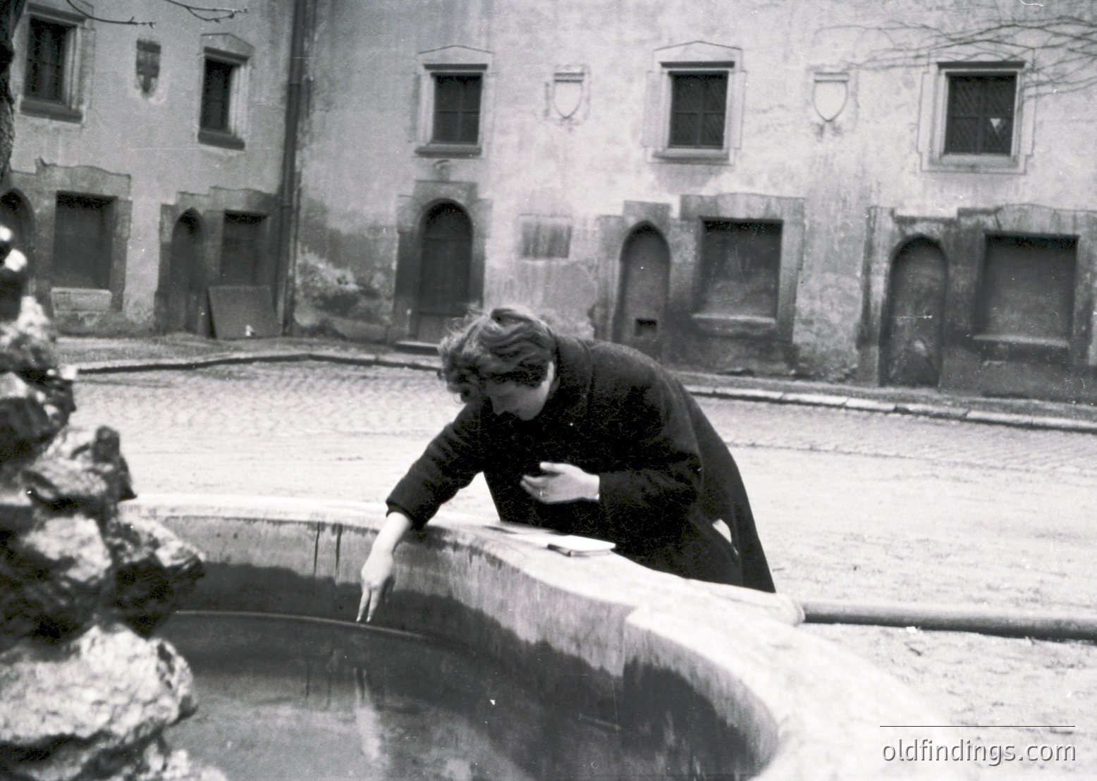 Black-and-white courtyard scene featuring a person in winter attire cleaning a stone fountain basin, surrounded by aged stone walls with arched doorways. Likely 1950s–1970s European urban setting.