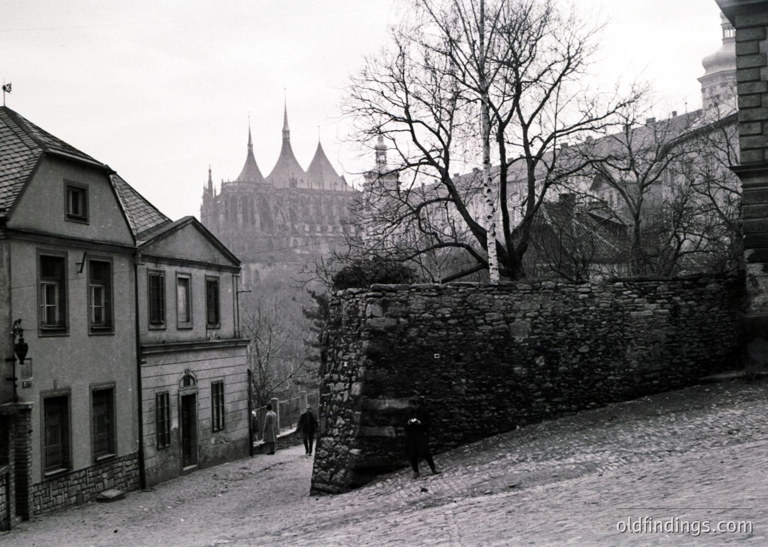 Black-and-white street scene in **Bucharest, Romania**, featuring the iconic **Patriarchal Cathedral** in the background. Cobblestone road flanked by historic buildings with wrought-iron street lamps and a vintage car parked near a stone wall. Mid-20th century urban architecture with Gothic Revival influences.