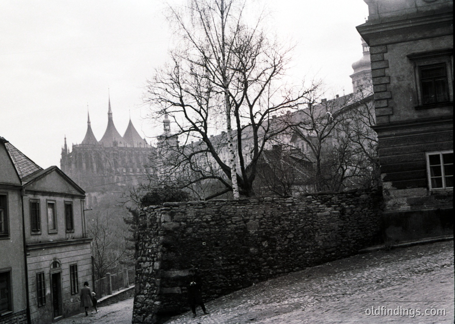Black-and-white street scene featuring Gothic spires of **Bratislava Castle** in the background, framed by leafless trees and cobblestone streets. Stone wall and residential buildings with arched windows dominate the foreground. Likely **1950s–1960s** Slovakia.